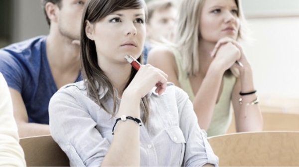 Mehrere junge Erwachsene sitzen aufmerksam nebeneinander in einem Seminarraum. Im Vordergrund hält eine Frau mit braunen Haaren einen Stift ans Kinn und blickt nachdenklich nach vorn, während andere ebenfalls konzentriert zuhören.
