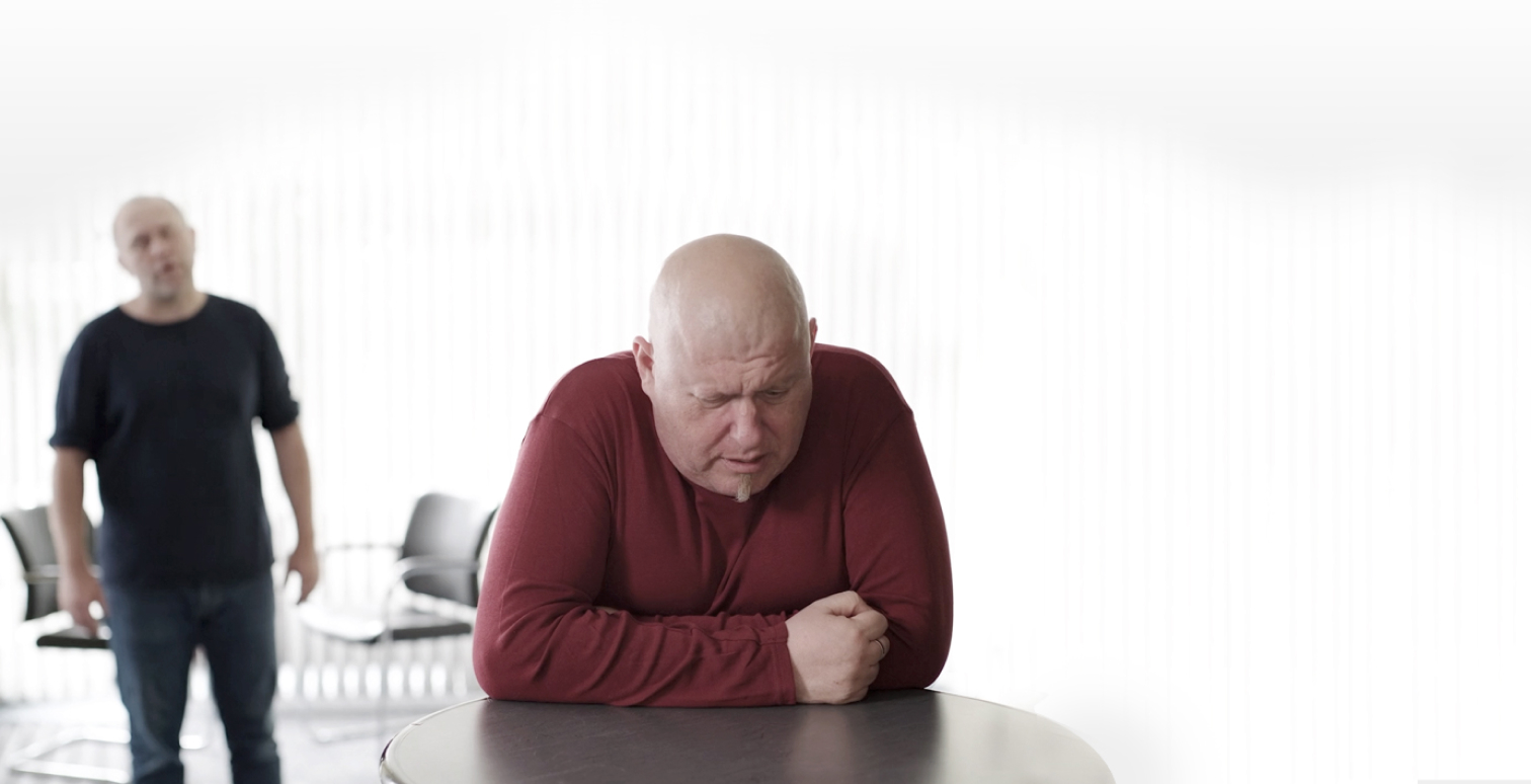 Ein glatzköpfiger Mann in rotem Shirt sitzt mit gesenktem Blick und verschränkten Armen an einem Tisch und wirkt bedrückt oder nachdenklich. Im Hintergrund steht ein weiterer Mann in dunkler Kleidung, leicht unscharf, in einem hellen Raum mit leeren Stühlen
