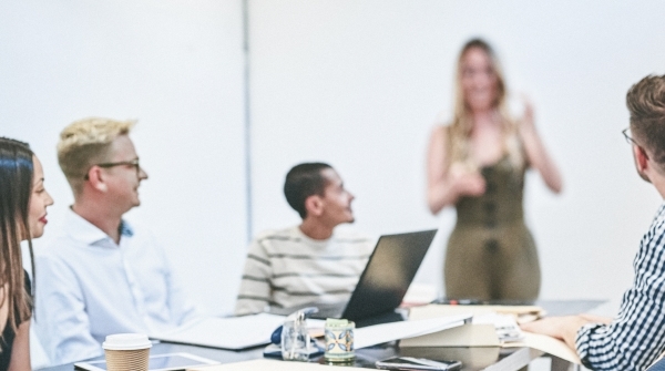 Fünf Personen in einem modernen Besprechungsraum: Vier sitzen mit Laptops und Getränken an einem Tisch und schauen lächelnd auf eine Frau, die vorne steht und spricht. Die Atmosphäre wirkt locker und kommunikativ